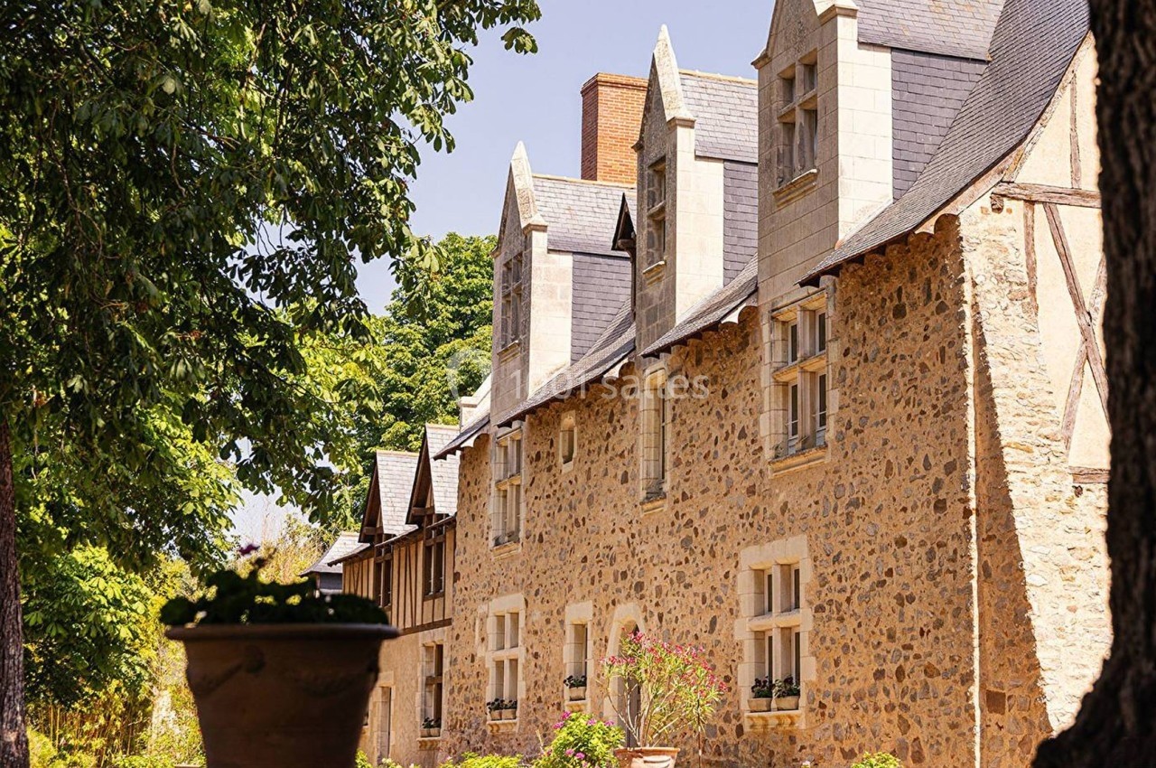 Façade en pierre d'une maison traditionnelle avec fenêtres à lucarnes, entourée d'arbres et de végétation.