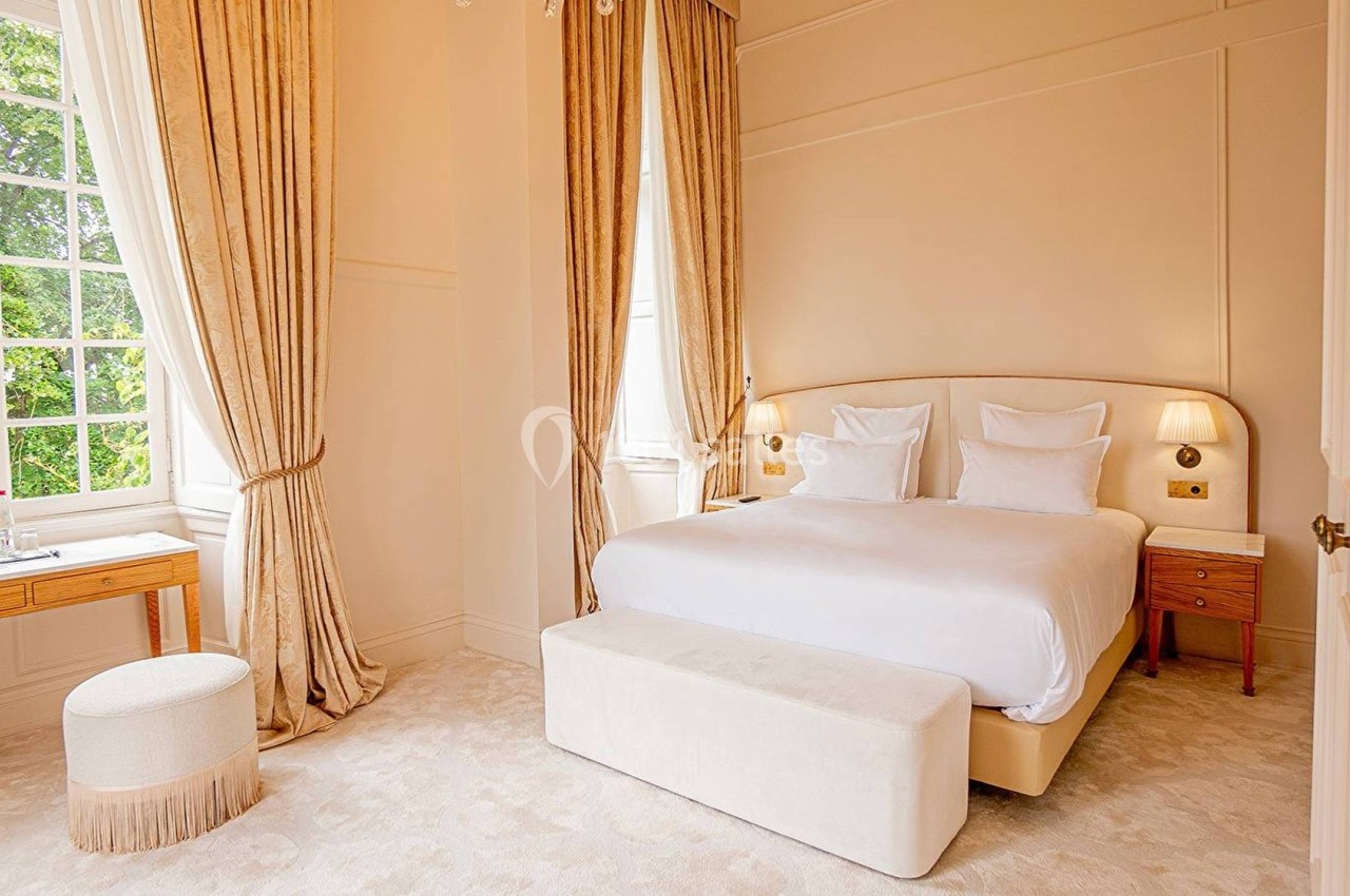 Chambre lumineuse avec un grand lit, des rideaux beiges, un banc au pied du lit et une fenêtre donnant sur des arbres.