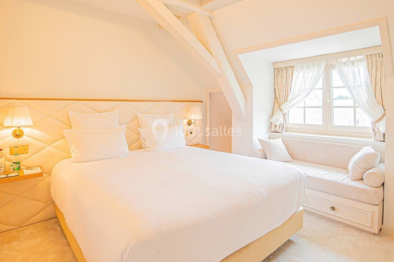 Chambre lumineuse avec lit double, tête de lit capitonnée, lampes murales et coin salon près d'une fenêtre.