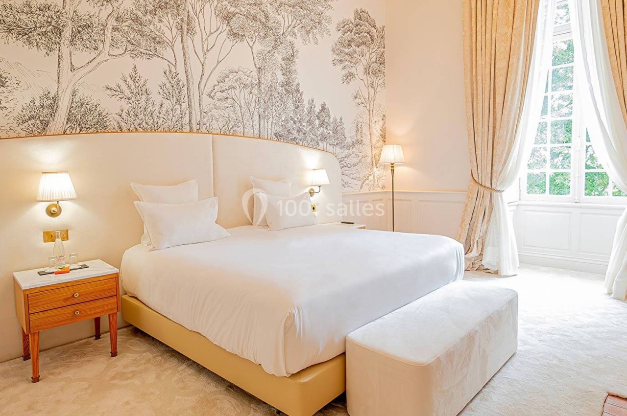 Chambre lumineuse avec lit double, tête de lit courbée, murs décorés de motifs naturels et grandes fenêtres avec rideaux.