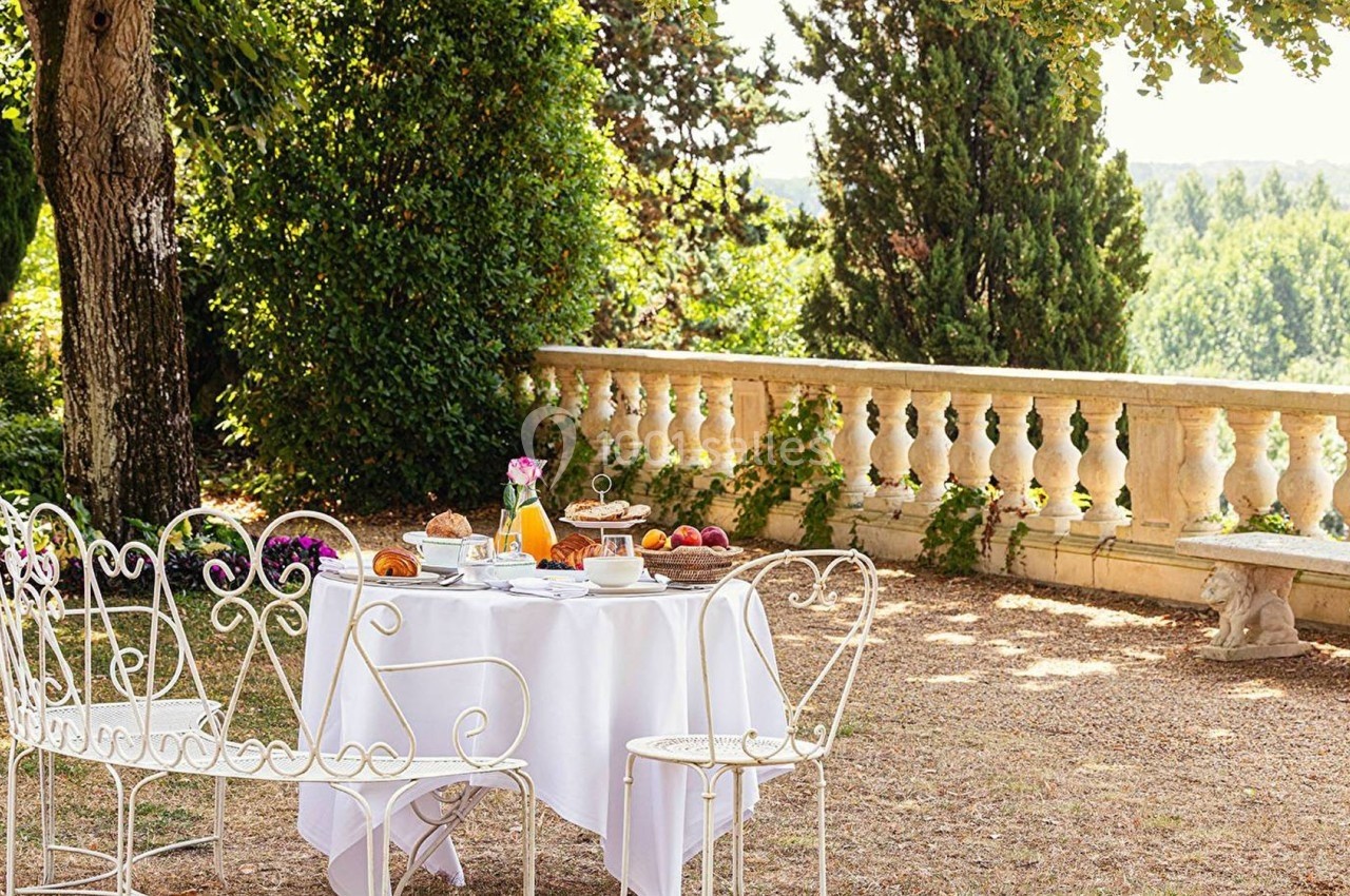 Table de jardin dressée avec petit-déjeuner, entourée de chaises en fer forgé, sous des arbres près d'une balustrade.
