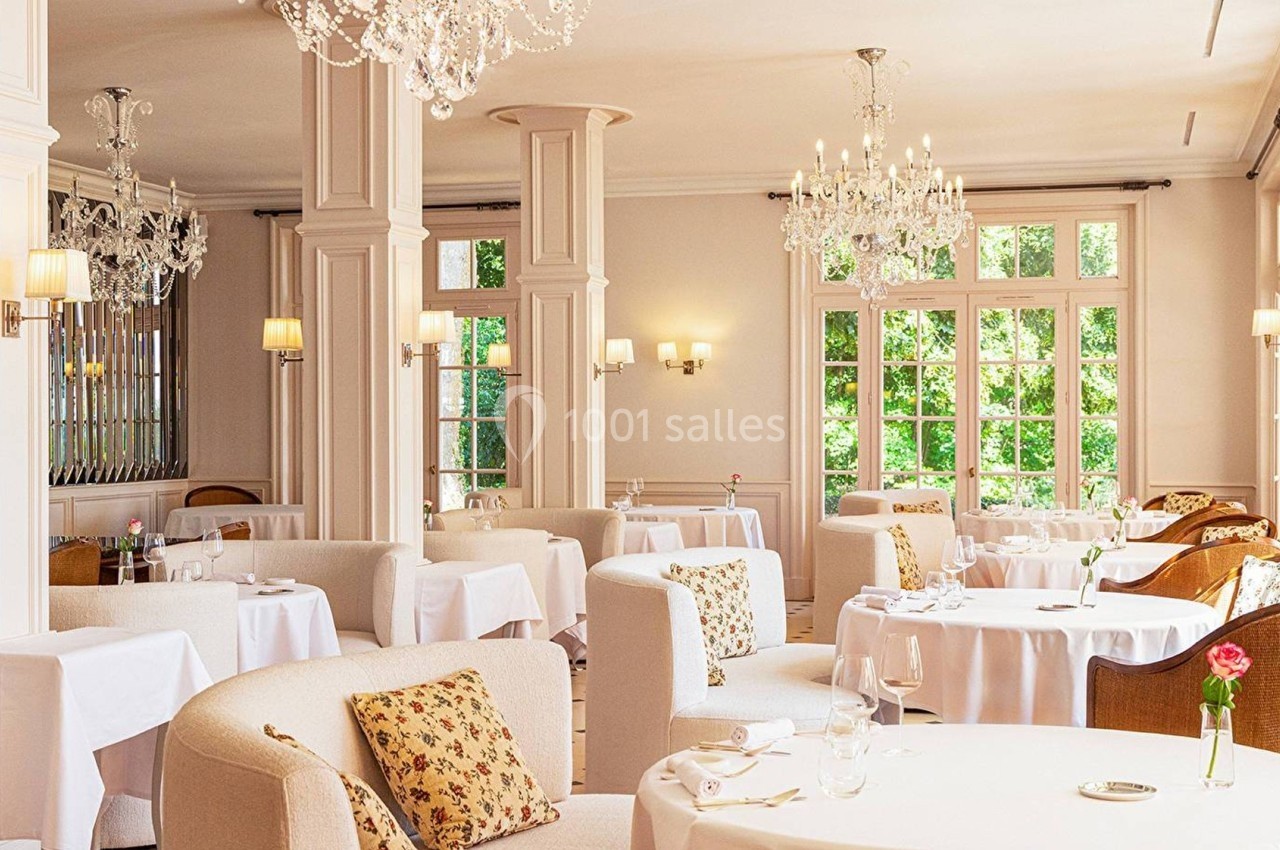 Salle de restaurant lumineuse avec tables dressées, fauteuils confortables et lustres en cristal.