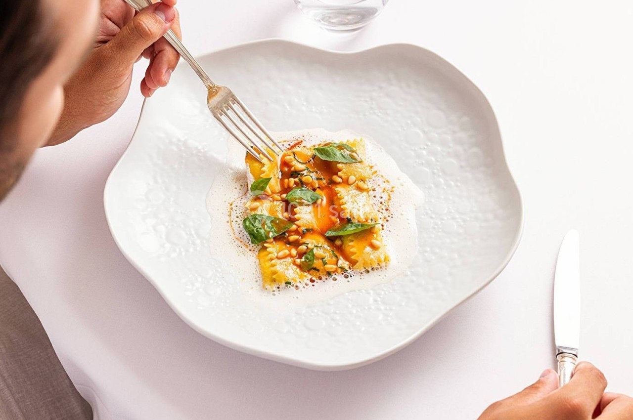 Assiette de raviolis garnis de sauce, basilic et pignons, tenue par une main avec une fourchette, sur une table blanche.