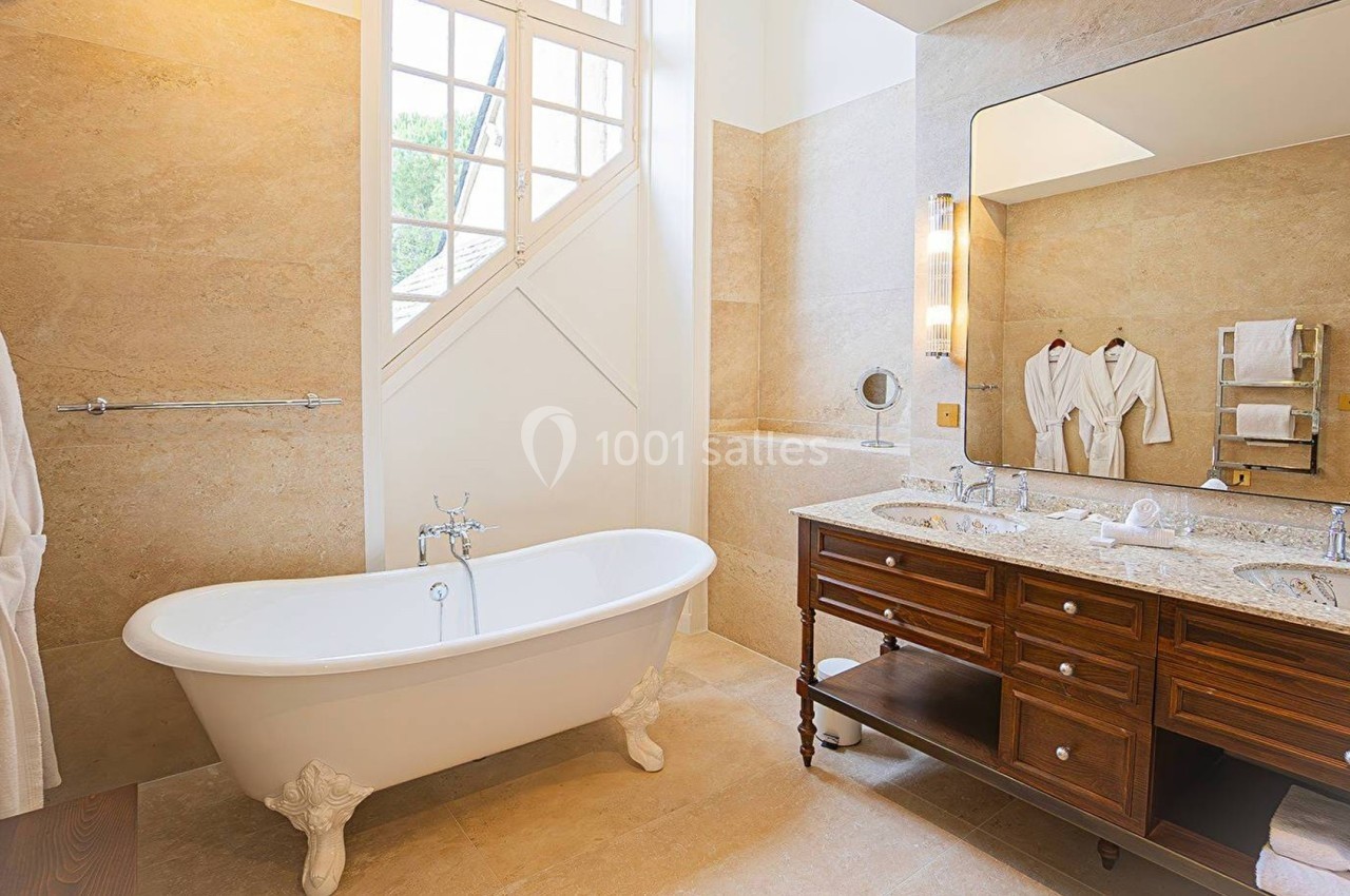 Salle de bain lumineuse avec baignoire sur pieds, double vasque en bois et miroirs, murs en pierre beige.
