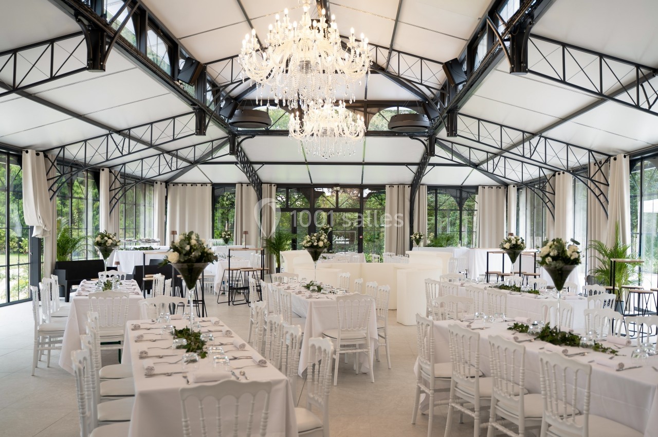Salle de réception lumineuse avec tables décorées, chaises blanches et un grand lustre central sous une verrière.