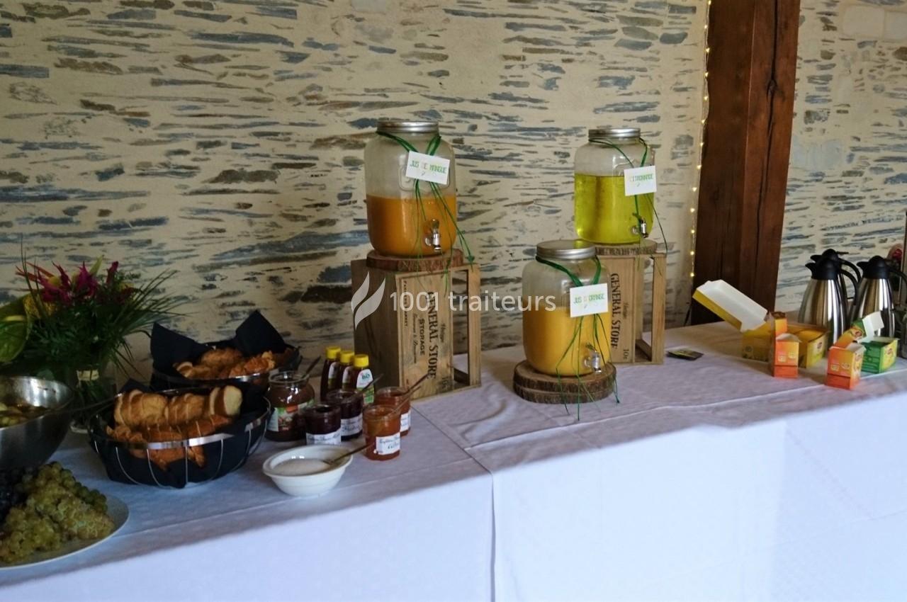 Buffet avec jus en distributeurs, viennoiseries, fruits, confitures et boissons chaudes sur une table blanche.