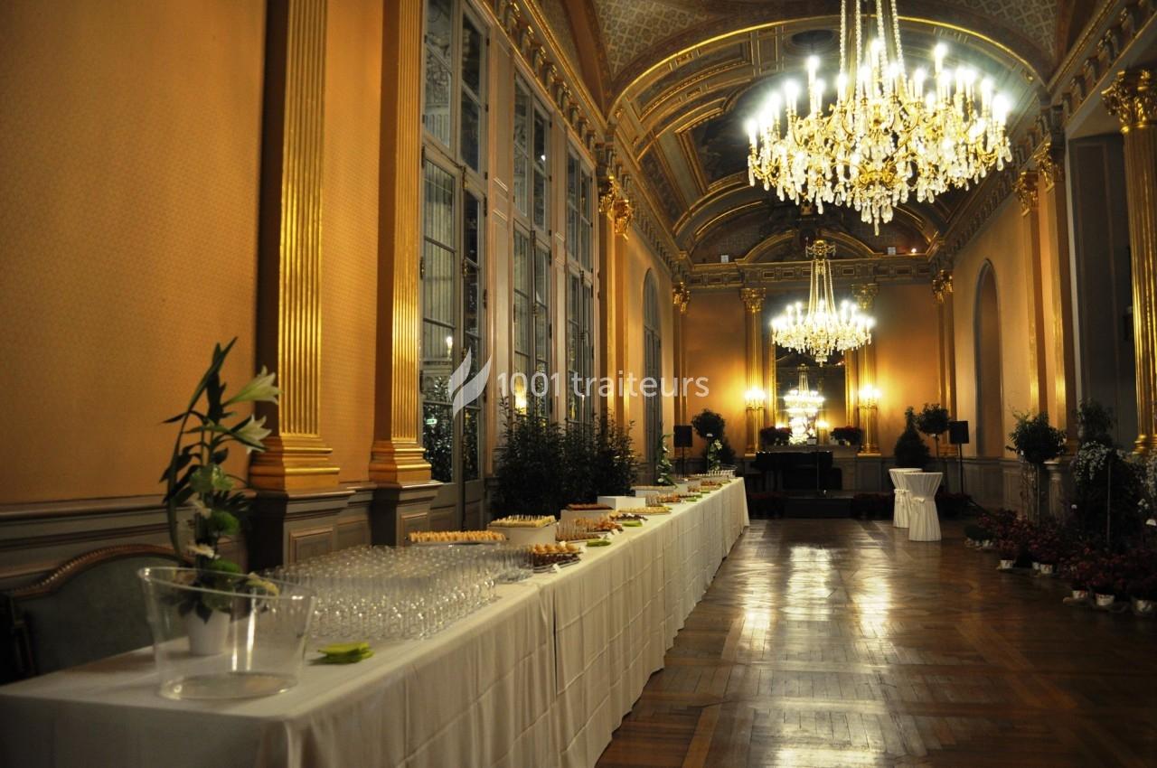 Salle élégante avec hauts plafonds, lustres en cristal, grandes fenêtres et tables dressées pour un buffet.