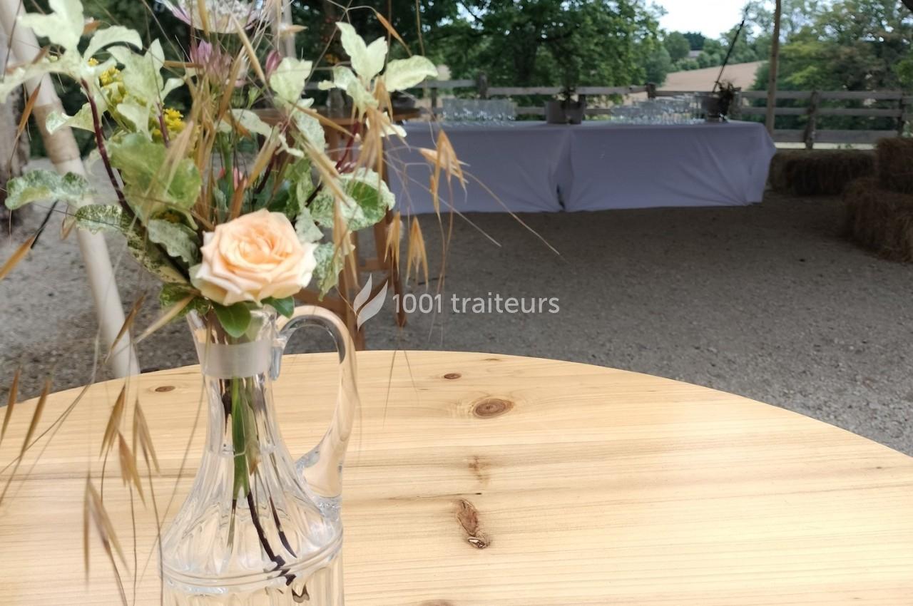 Vase en verre avec des fleurs séchées et une rose, posé sur une table en bois dans un espace extérieur avec des tables…