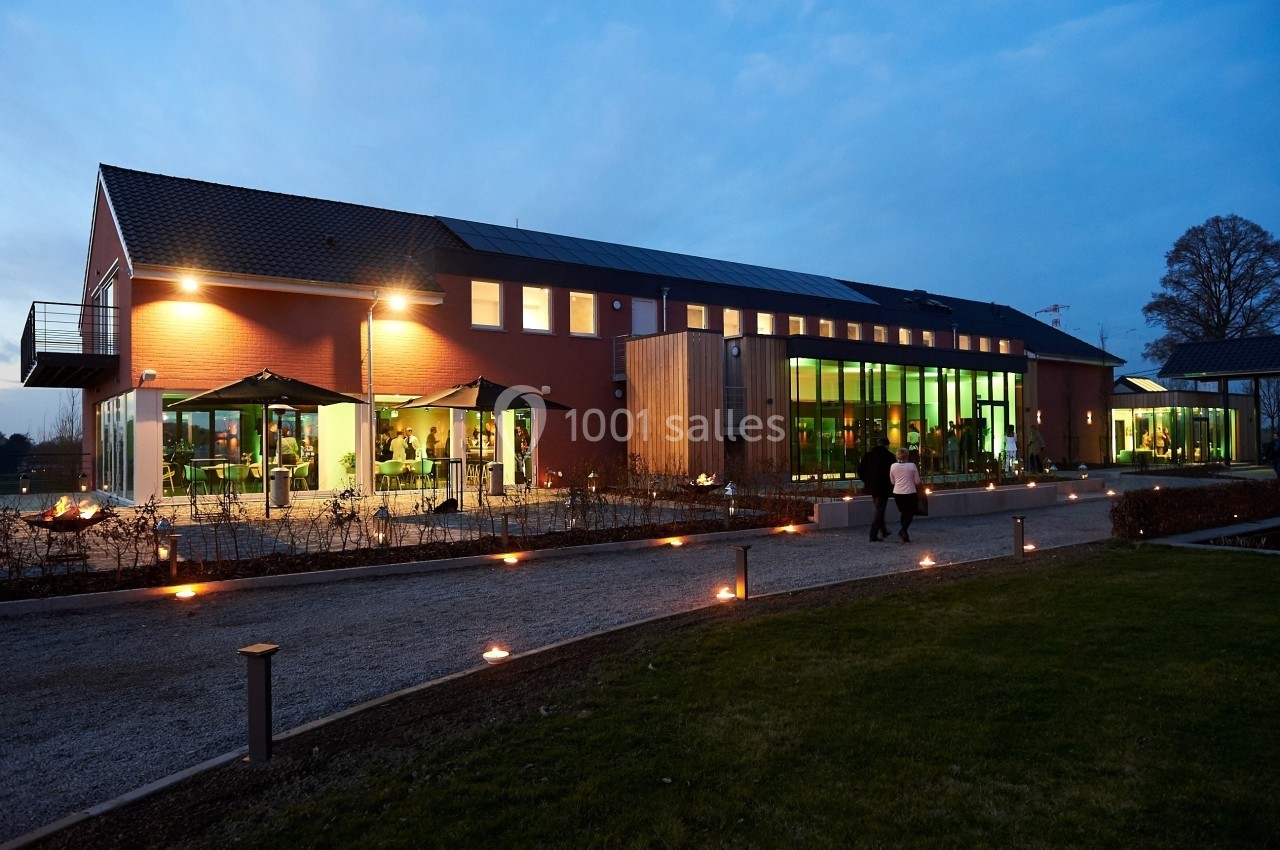 Bâtiment moderne éclairé en soirée, avec de grandes baies vitrées et une allée bordée de lumières au sol.
