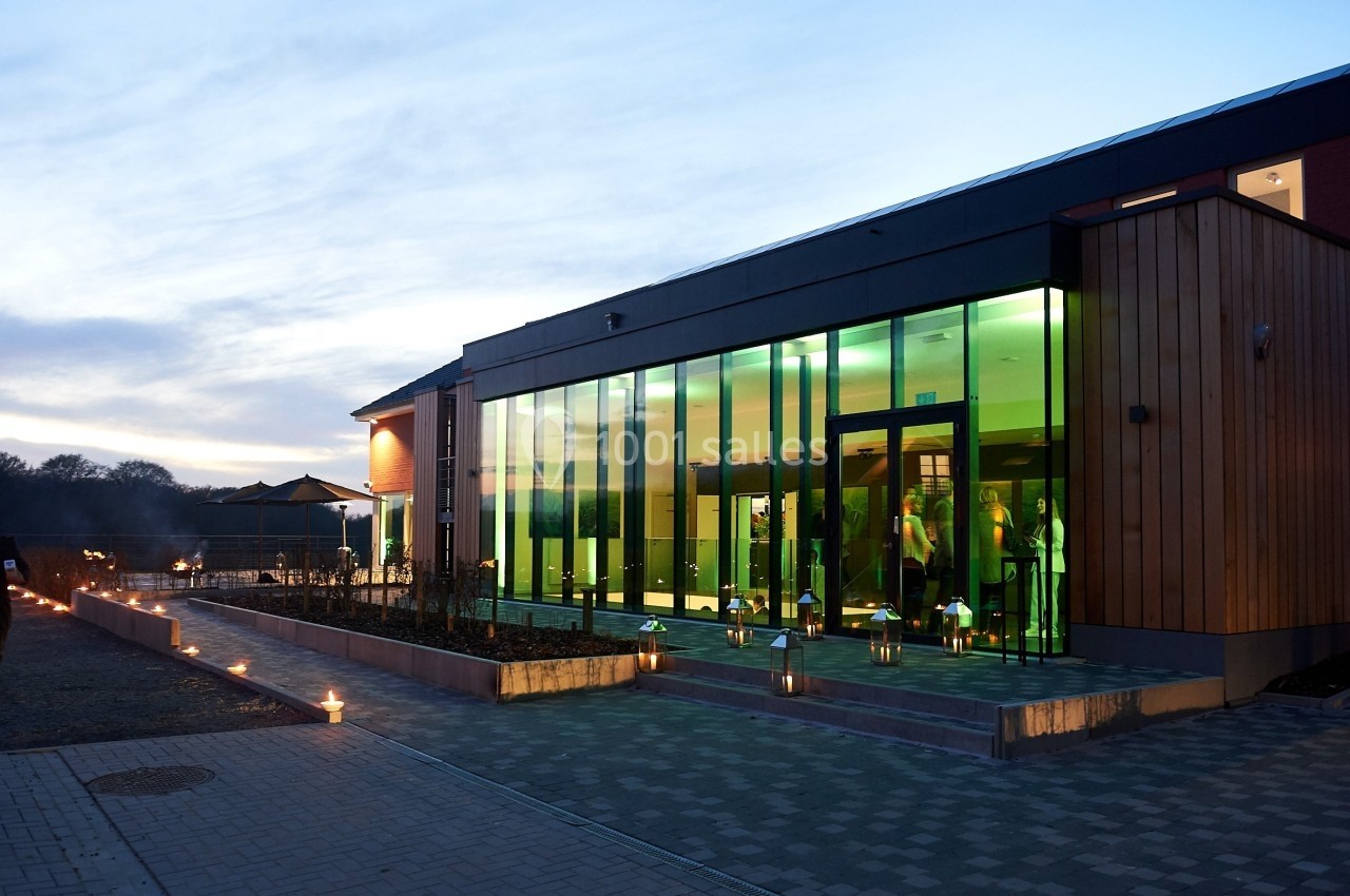 Façade d'un bâtiment moderne avec de grandes baies vitrées éclairées, entouré de bougies au crépuscule.
