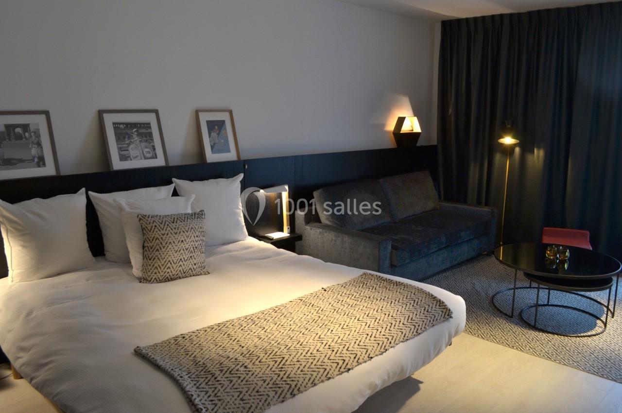 Chambre moderne avec lit double, canapé, table basse et éclairage tamisé, décorée de cadres et rideaux sombres.