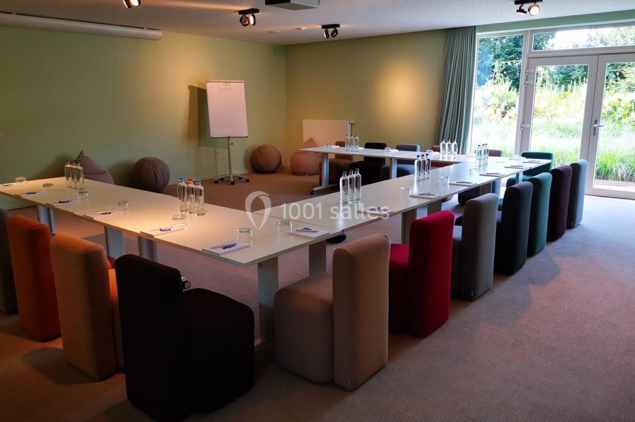 Salle de réunion lumineuse avec tables en U, chaises colorées, tableau blanc et vue sur un jardin.