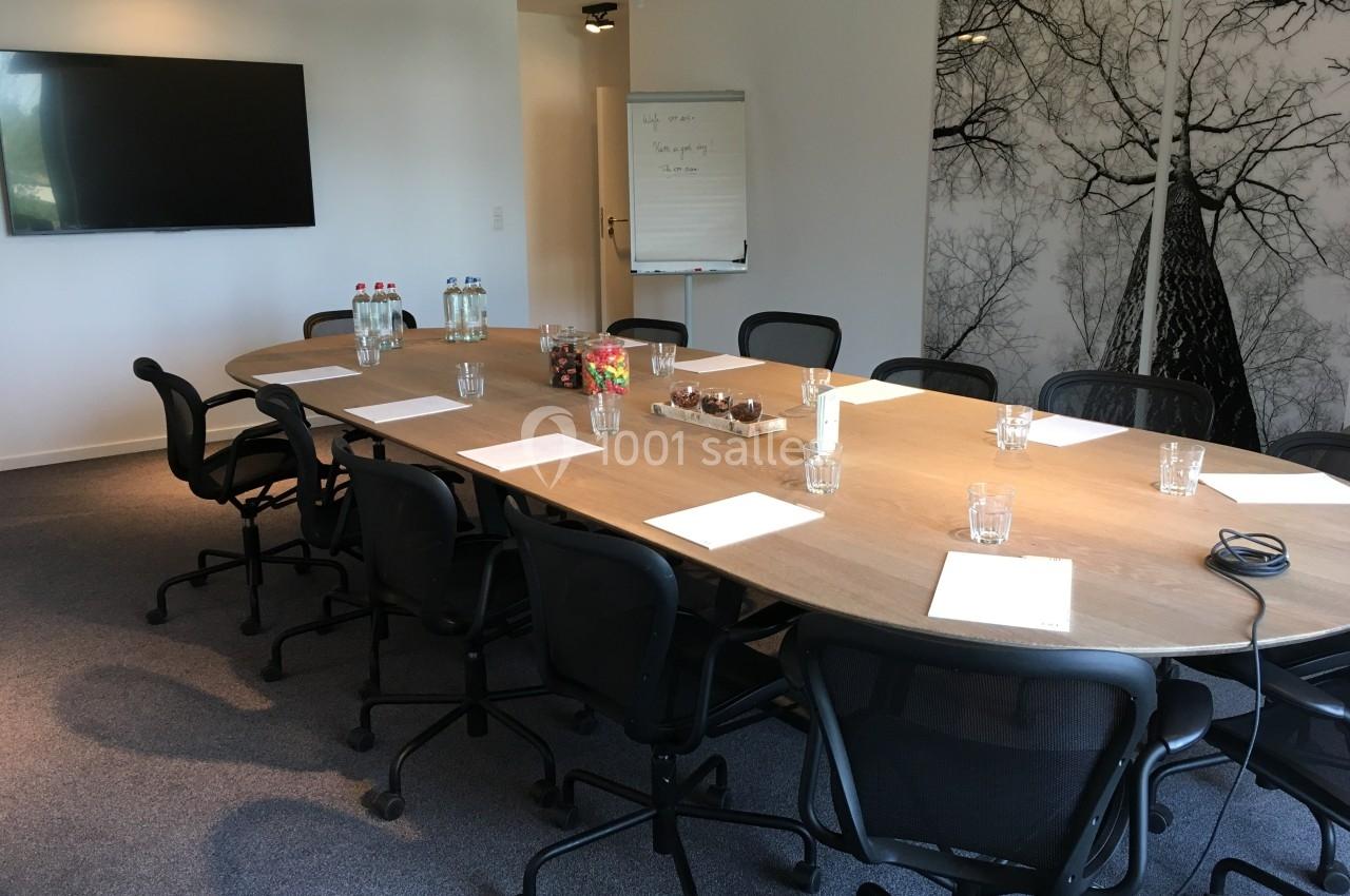 Salle de réunion avec une grande table en bois, chaises noires, écran mural, paperboard et fournitures disposées.