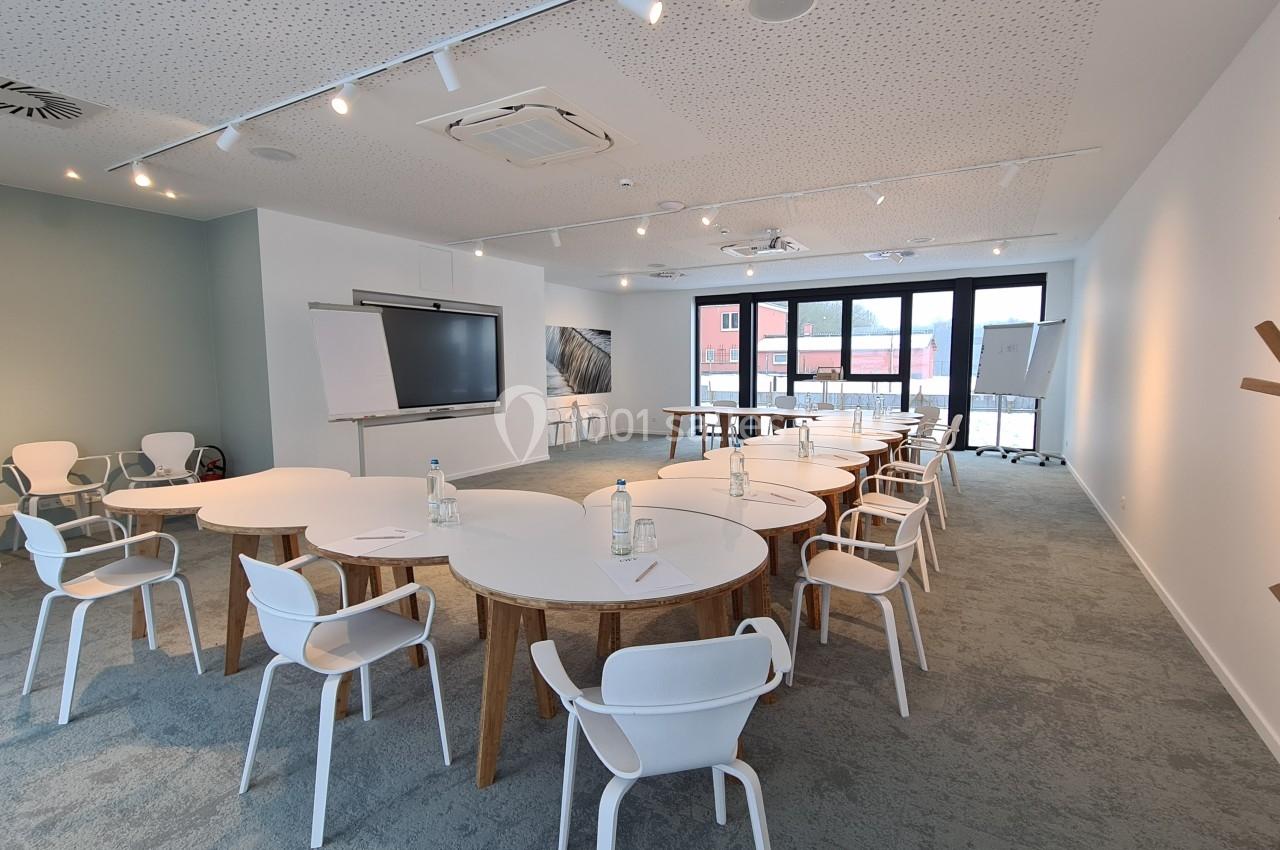 Salle de réunion lumineuse avec des tables rondes, des chaises blanches, un tableau blanc et un écran mural.