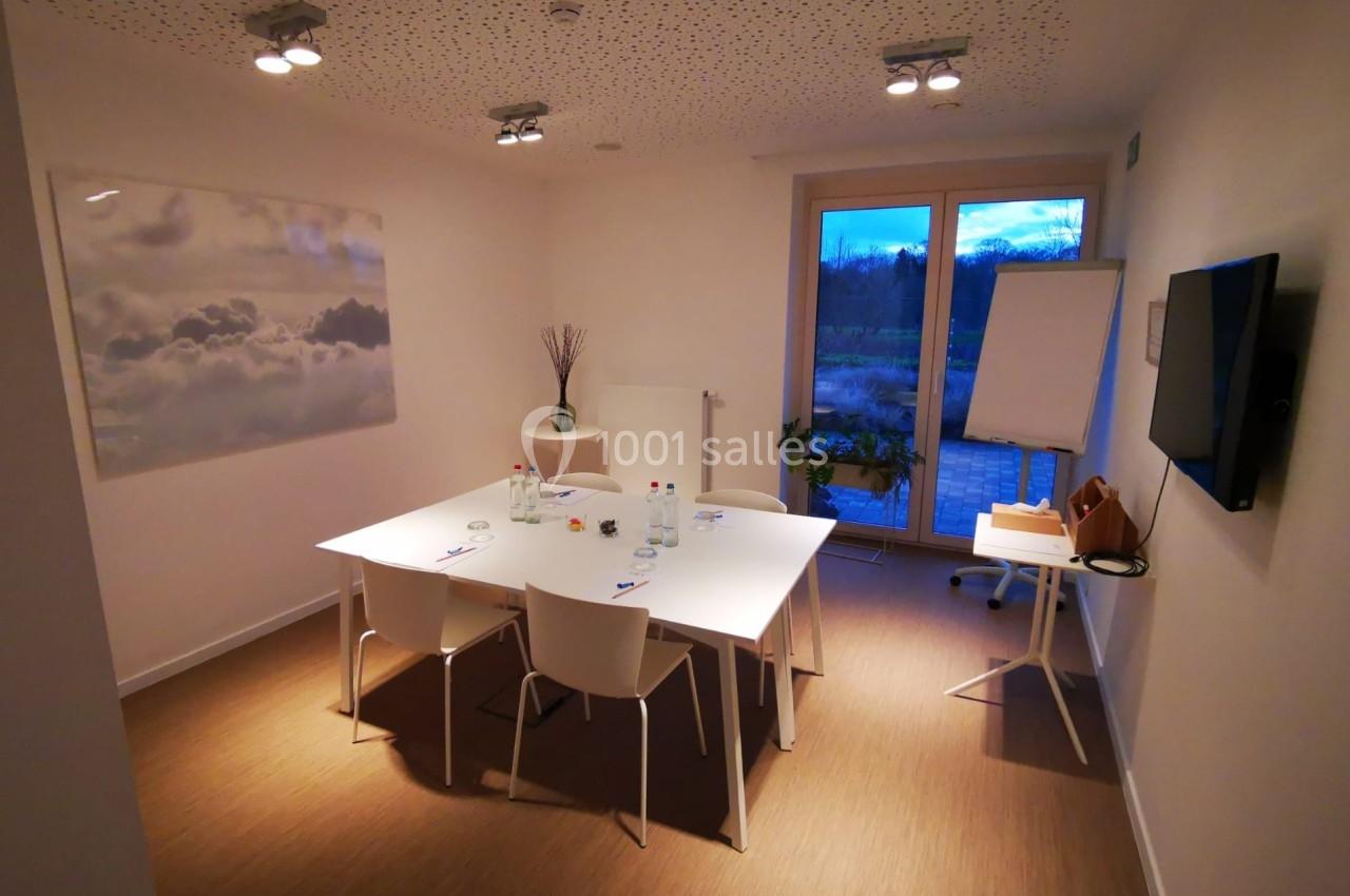 Salle de réunion lumineuse avec table blanche, chaises, tableau de conférence et vue sur l'extérieur en soirée.