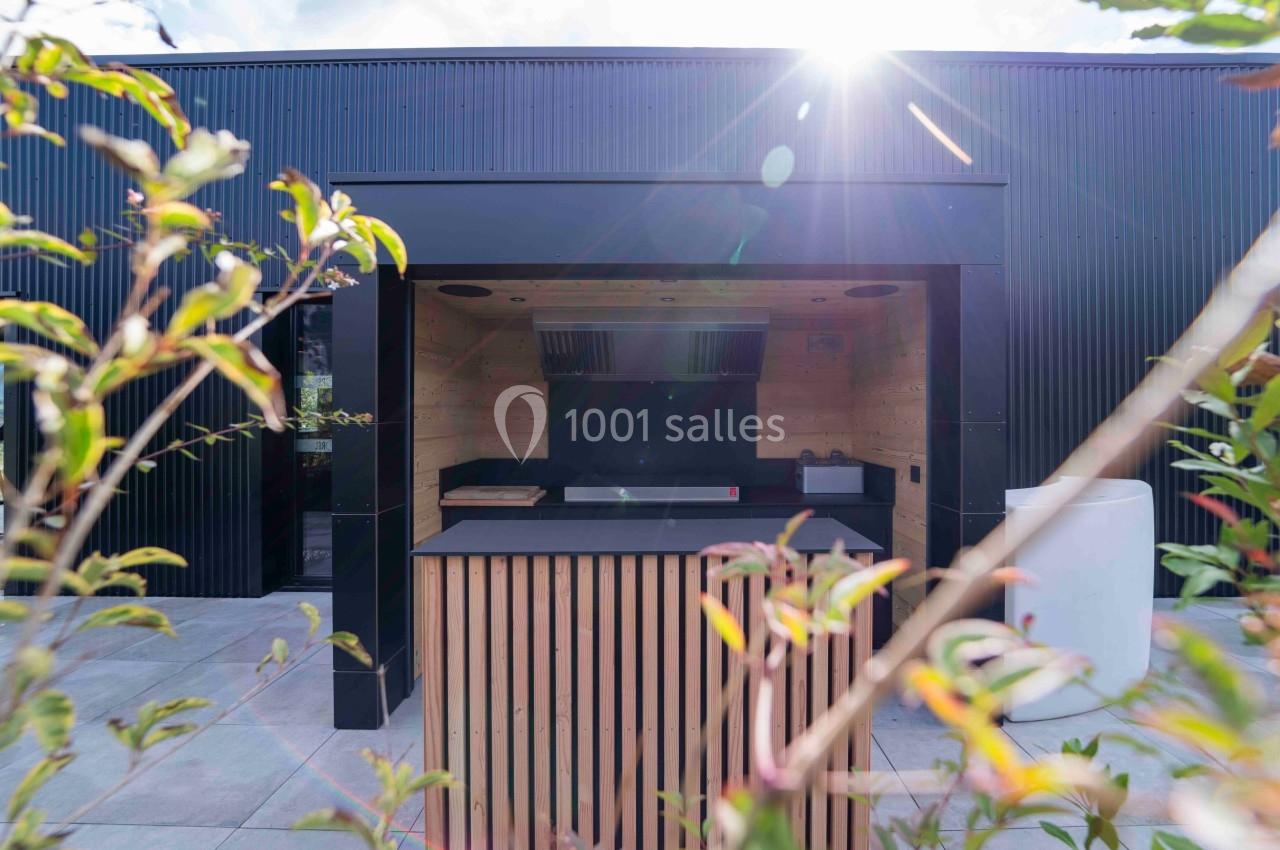 Cuisine extérieure moderne avec comptoir en bois, abri noir et lumière naturelle filtrée par des plantes au premier plan.