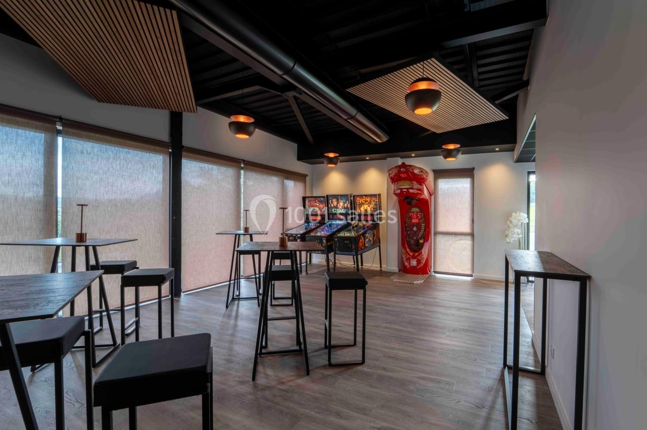 Salle moderne avec tables hautes, chaises noires et jeux d'arcade alignés près d'une baie vitrée avec stores baissés.