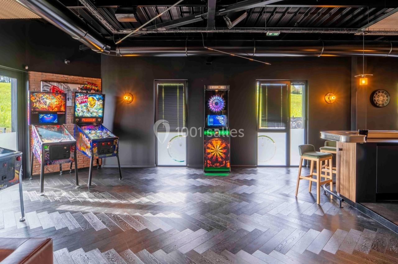 Salle de jeux avec flippers, cible de fléchettes électronique et mobilier moderne sur un sol en parquet sombre.