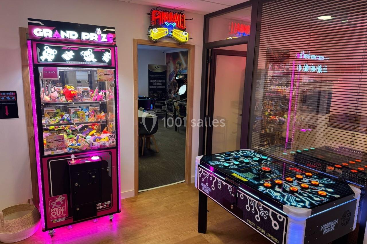 Salle avec une machine à pinces lumineuse, une table de jeu rétro et une enseigne ’Pinball’ au-dessus d'une porte.