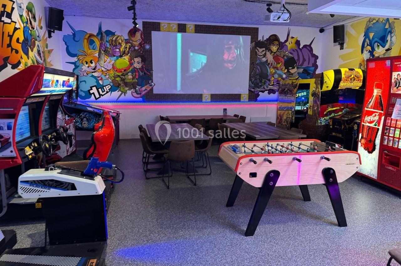 Salle d'arcade avec baby-foot, bornes de jeux, tables, fresques murales colorées et écran projetant un film.