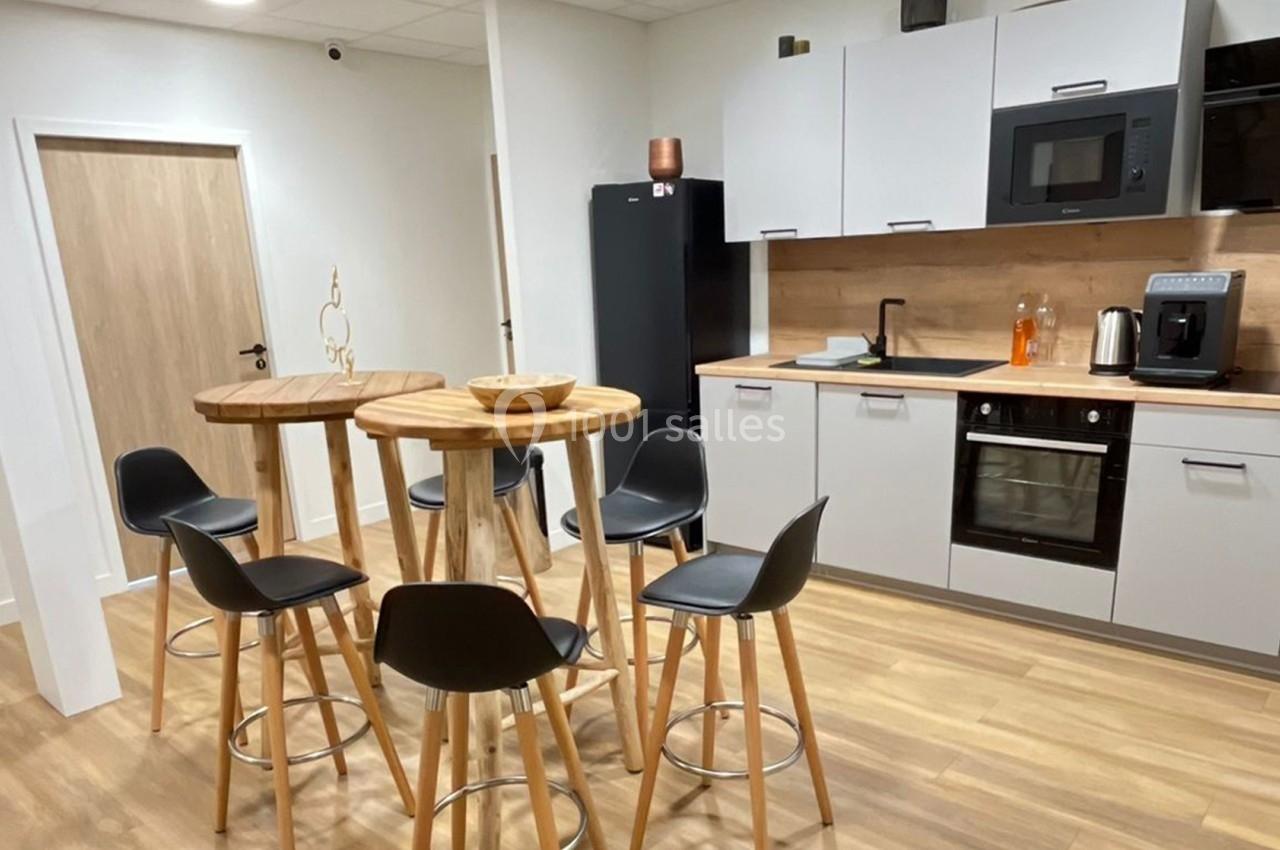 Cuisine moderne avec plancher en bois, tables hautes, chaises noires, réfrigérateur, micro-ondes et cafetière.