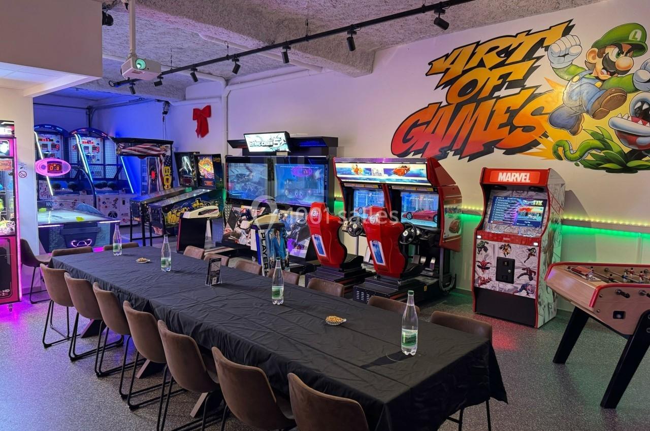 Salle d'arcade avec des bornes de jeux, flippers et une grande table noire entourée de chaises.