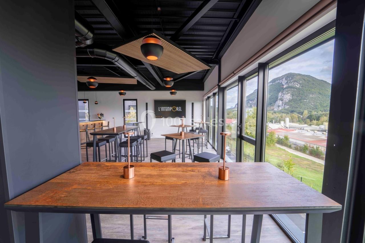 Salle moderne avec grandes baies vitrées, tables en bois, chaises hautes et vue sur un paysage montagneux.