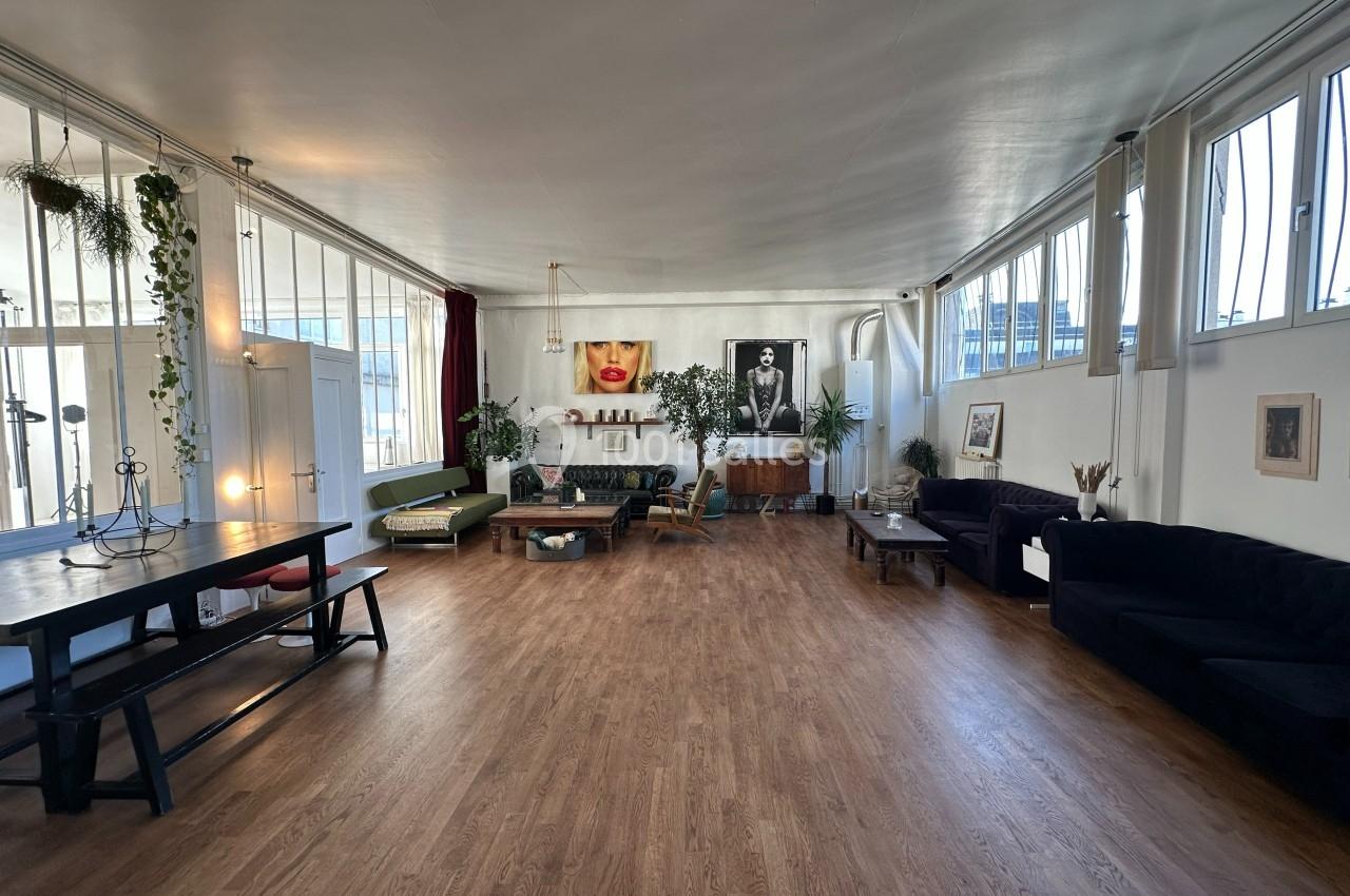 Salle spacieuse avec parquet, mobilier éclectique, plantes suspendues et grandes fenêtres laissant entrer la lumière…