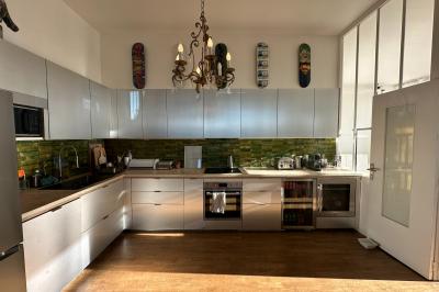 Salle lumineuse avec parquet, canapés sombres, plantes, décorations murales et grandes fenêtres laissant entrer la lumière.