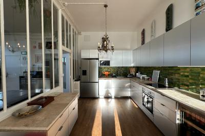 Salle lumineuse avec parquet, canapés sombres, plantes, décorations murales et grandes fenêtres laissant entrer la lumière.