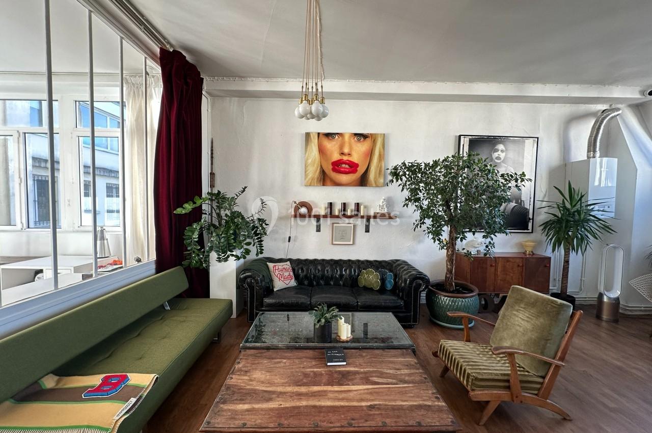 Salon lumineux avec canapé vert, table basse en bois, plantes, œuvres d'art au mur et grande verrière.