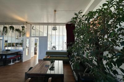 Salle lumineuse avec parquet, canapés sombres, plantes, décorations murales et grandes fenêtres laissant entrer la lumière.