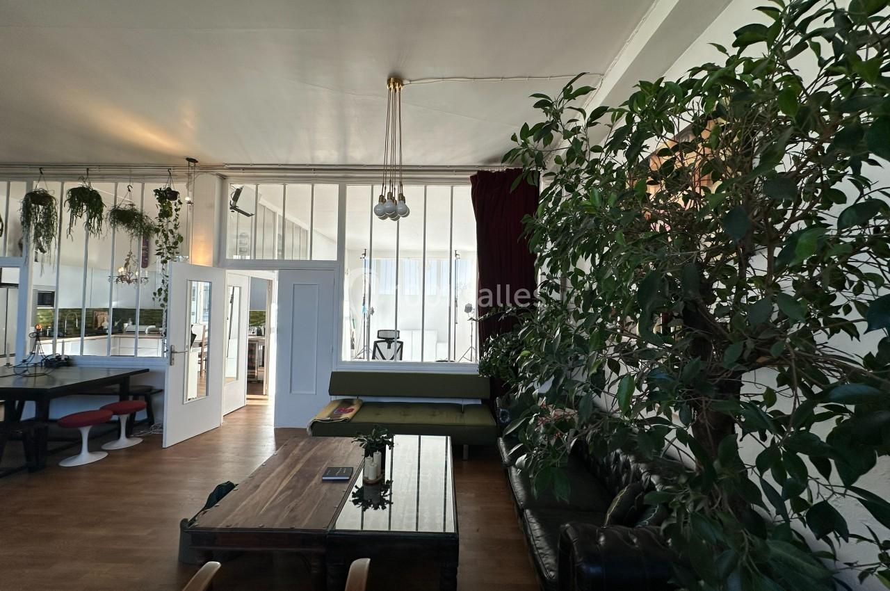 Salon lumineux avec de grandes baies vitrées, mobilier en bois et plantes vertes décoratives.