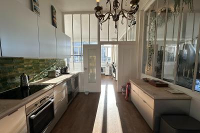 Salle lumineuse avec parquet, canapés sombres, plantes, décorations murales et grandes fenêtres laissant entrer la lumière.