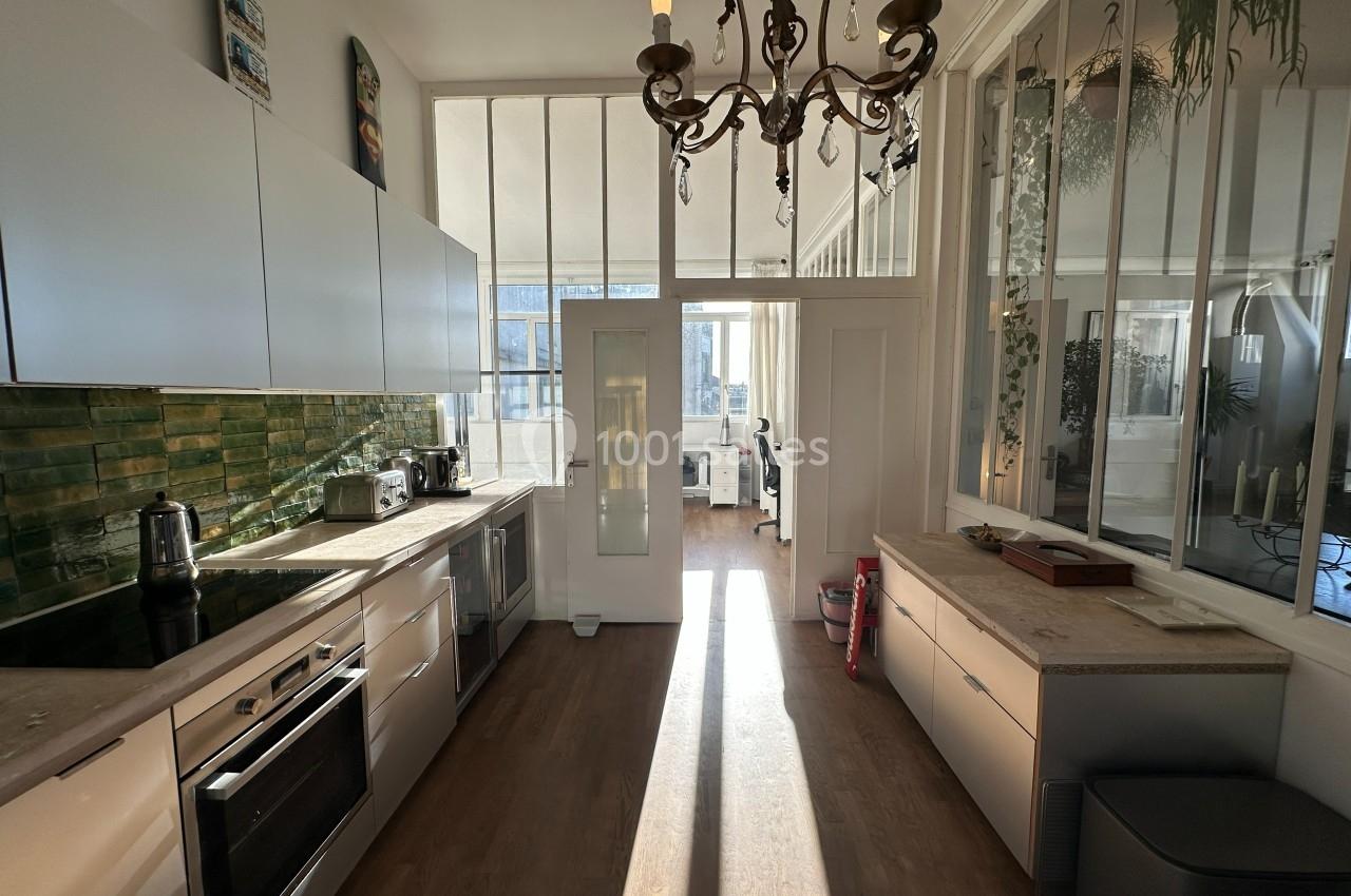 Cuisine lumineuse avec mobilier moderne, crédence verte, et vue sur un espace de travail à travers des verrières.