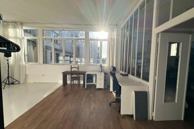 Salle lumineuse avec parquet, canapés sombres, plantes, décorations murales et grandes fenêtres laissant entrer la lumière.