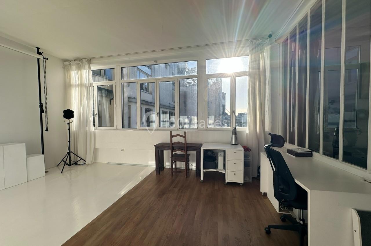 Salle lumineuse avec de grandes fenêtres, un bureau, une chaise, un éclairage studio et un sol en bois.