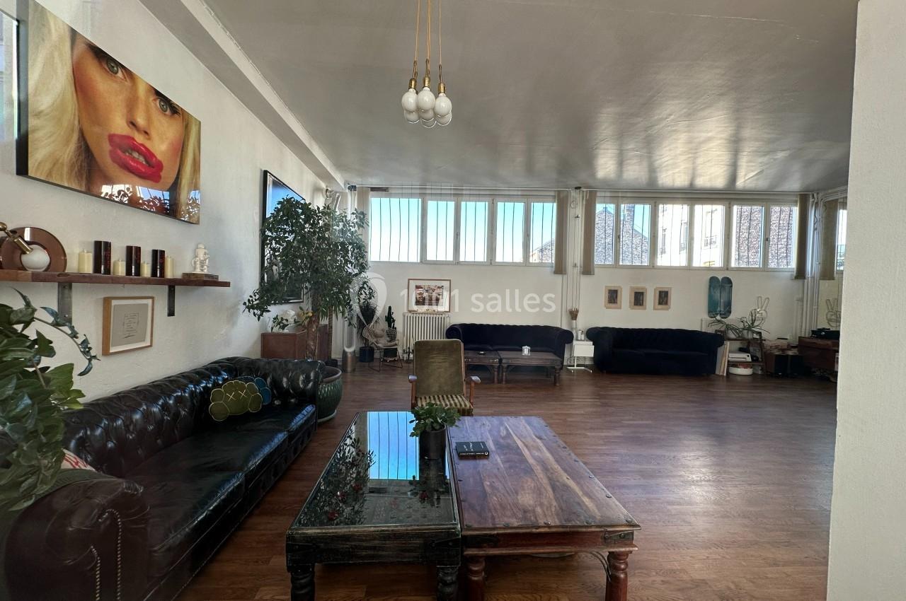 Salon spacieux avec parquet, canapés en cuir et velours, table basse en bois, plantes et grandes fenêtres laissant entrer la…