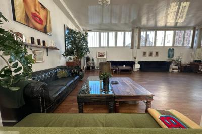 Salle lumineuse avec parquet, canapés sombres, plantes, décorations murales et grandes fenêtres laissant entrer la lumière.