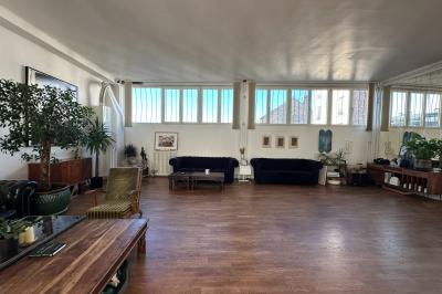 Salle lumineuse avec parquet, canapés sombres, plantes, décorations murales et grandes fenêtres laissant entrer la lumière.