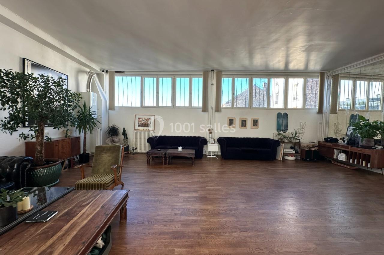 Salle spacieuse avec parquet, canapés, plantes et grandes fenêtres laissant entrer la lumière naturelle.