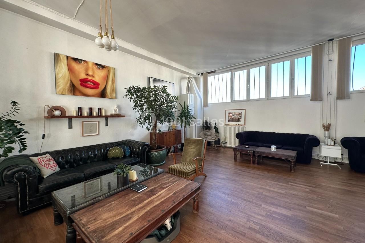 Salon spacieux avec canapés en cuir, tables en bois, plantes et grande fenêtre laissant entrer la lumière naturelle.