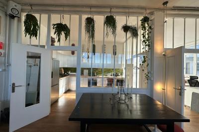 Salle lumineuse avec parquet, canapés sombres, plantes, décorations murales et grandes fenêtres laissant entrer la lumière.