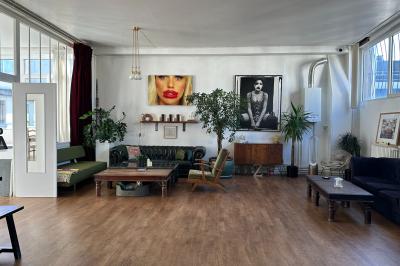 Salle lumineuse avec parquet, canapés sombres, plantes, décorations murales et grandes fenêtres laissant entrer la lumière.