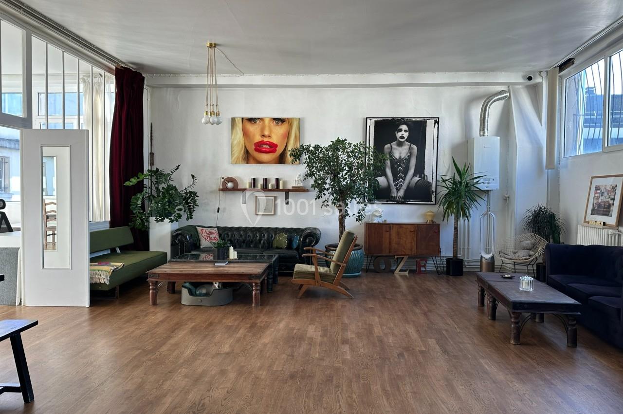 Salon lumineux avec parquet, canapés, plantes, tables basses et portraits accrochés sur un mur blanc.