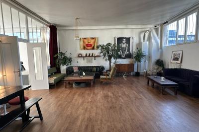 Salle lumineuse avec parquet, canapés sombres, plantes, décorations murales et grandes fenêtres laissant entrer la lumière.