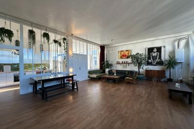 Salle lumineuse avec parquet, canapés sombres, plantes, décorations murales et grandes fenêtres laissant entrer la lumière.