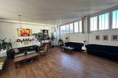 Salle lumineuse avec parquet, canapés sombres, plantes, décorations murales et grandes fenêtres laissant entrer la lumière.