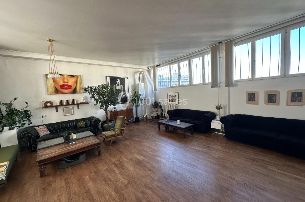 Salon spacieux avec parquet, canapés, plantes, tables basses et grandes fenêtres laissant entrer la lumière naturelle.