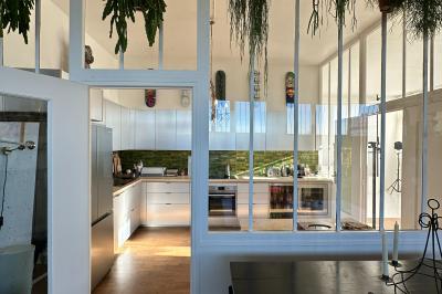 Salle lumineuse avec parquet, canapés sombres, plantes, décorations murales et grandes fenêtres laissant entrer la lumière.